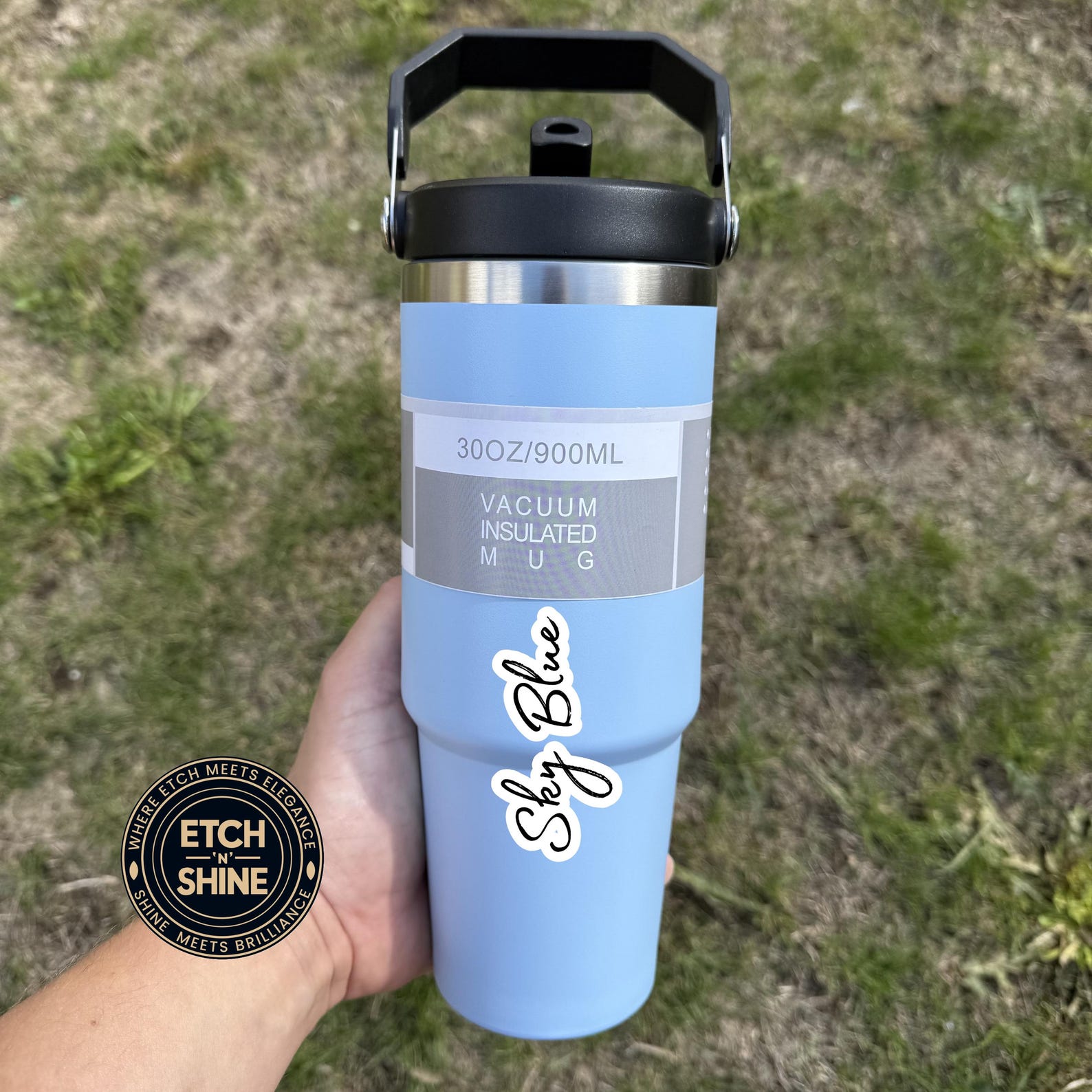 Light blue vacuum insulated mug with black lid held in a hand against a grassy background