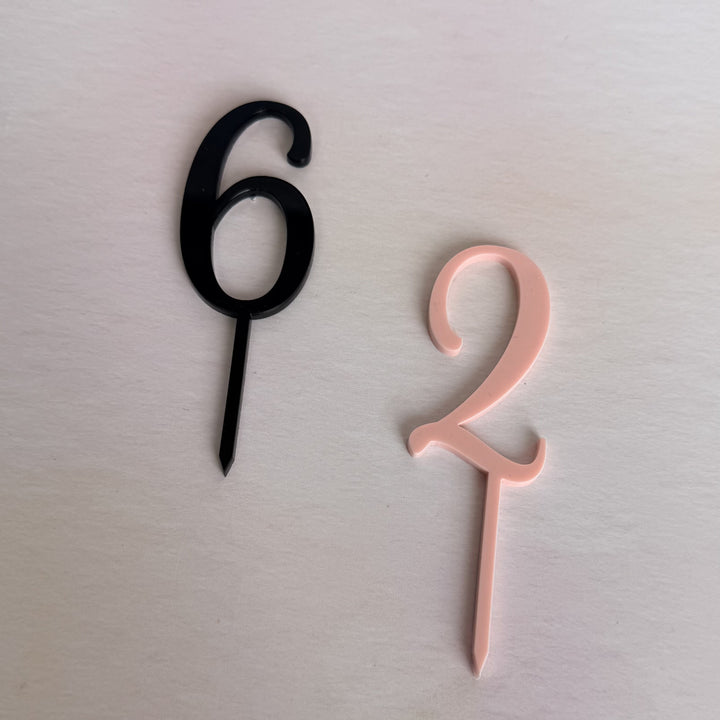 Two acrylic number cake toppers, one in matte black and one in light peach pink, displayed on a light beige surface.