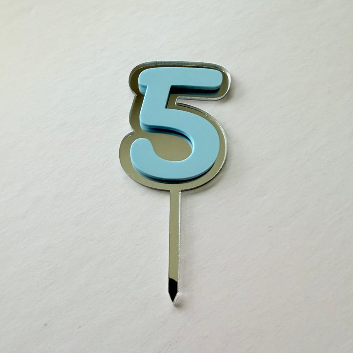 A single number 5 birthday candle with a light blue face and a silver metal outline and wick, resting on a plain off-white textured surface.