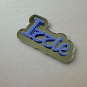 A small enamel pin or badge with the word 'Izzie' in blue script on a metallic gold cloud-shaped background, photographed on a plain light surface.