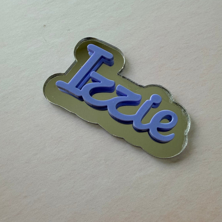 A small enamel pin or badge with the word 'Izzie' in blue script on a metallic gold cloud-shaped background, photographed on a plain light surface.