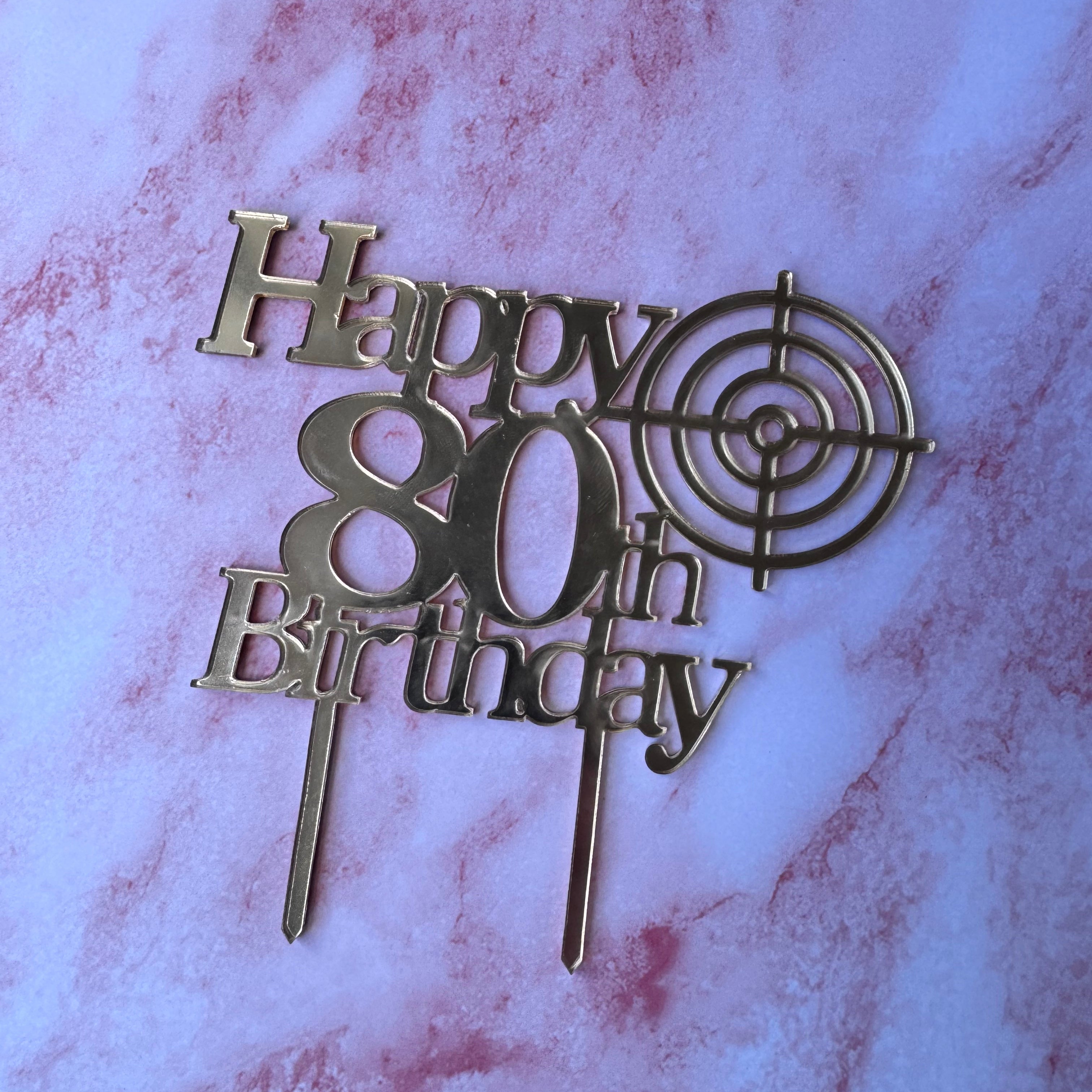 Gold metal 80th birthday cake topper with 'Happy 80th Birthday' text and a target design, displayed on a pink and white marble surface.