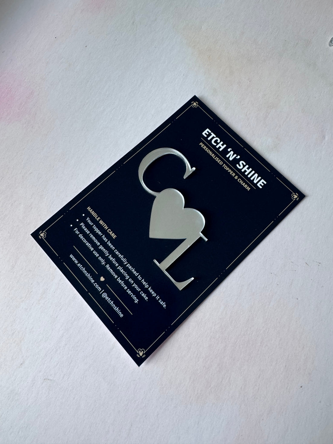 A silver metal heart-shaped letter etcher tool displayed on its black retail backing card, which reads 'ETCH 'N' SHINE'.