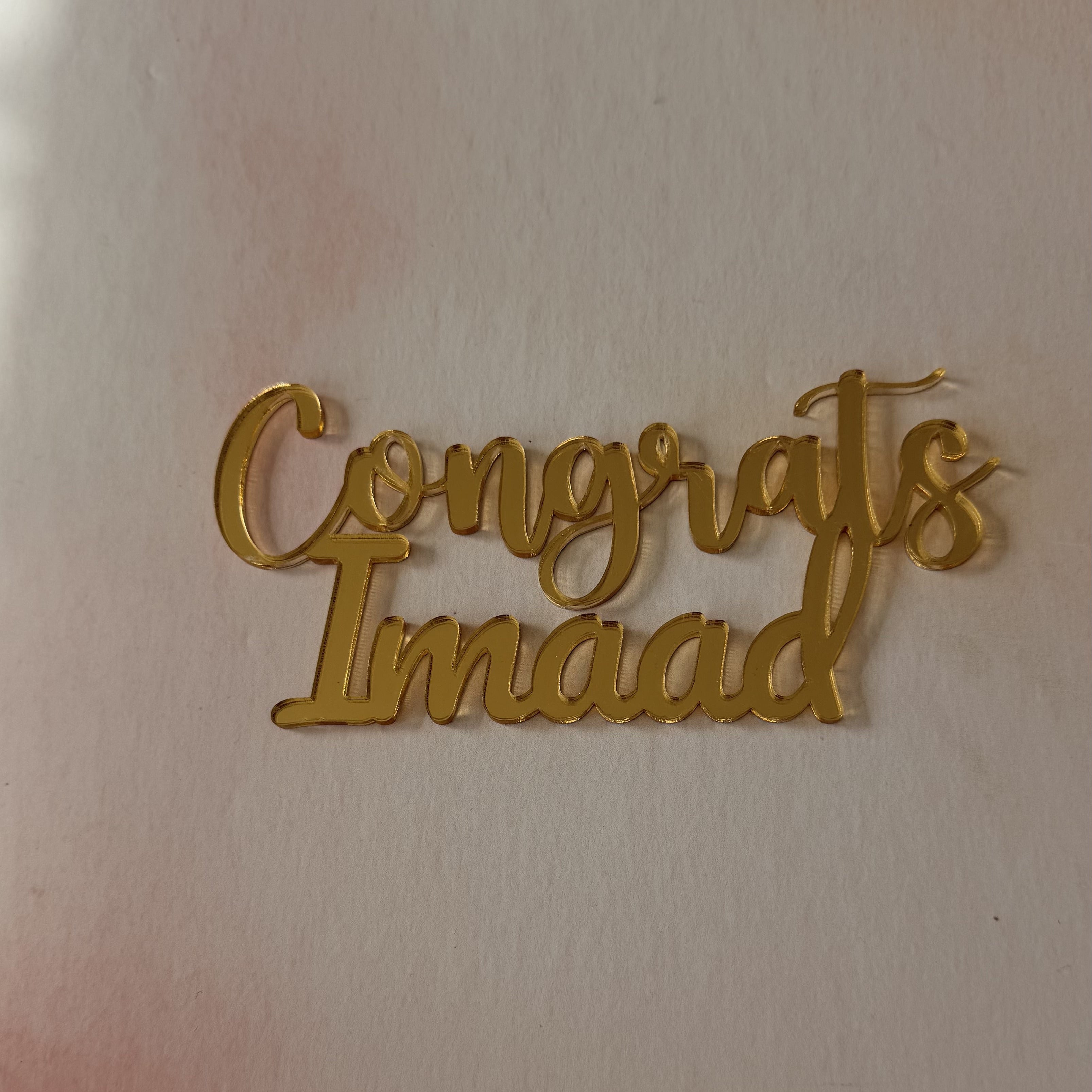 Gold metallic script lettering that reads 'Congrats Imaad' on a light beige background.