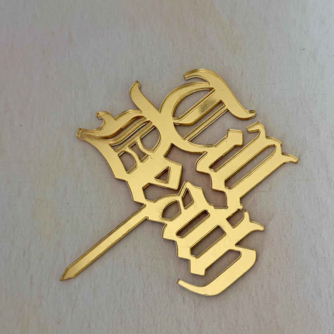 A gold metal gothic-style letter cake topper with a pointed stake, shown on a light beige textured background.