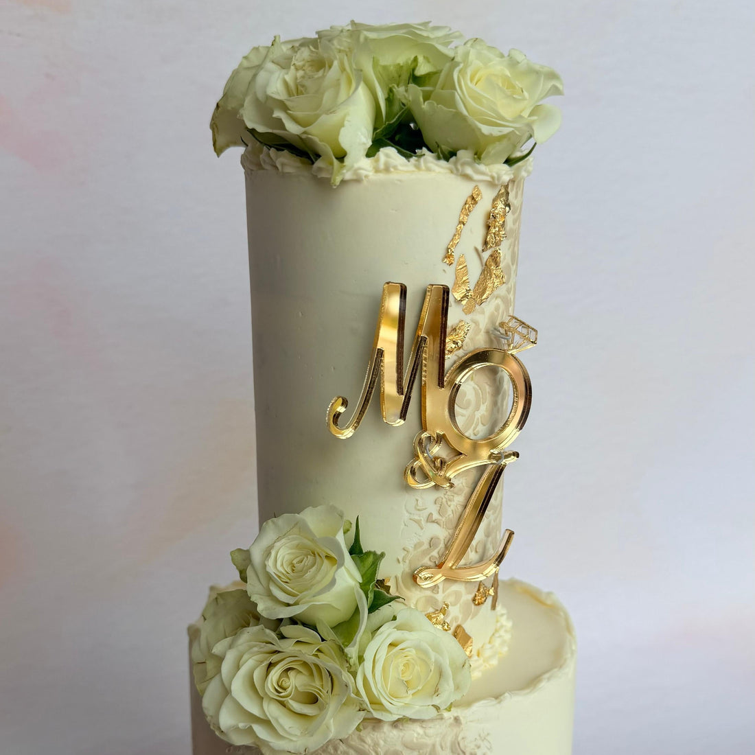 A two-tier custom wedding cake decorated with cream-colored roses, gold foil accents, and a gold monogram topper reading 'MBZ'.