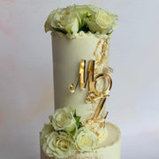 A two-tier custom wedding cake decorated with cream-colored roses, gold foil accents, and a gold monogram topper reading 'MBZ'.