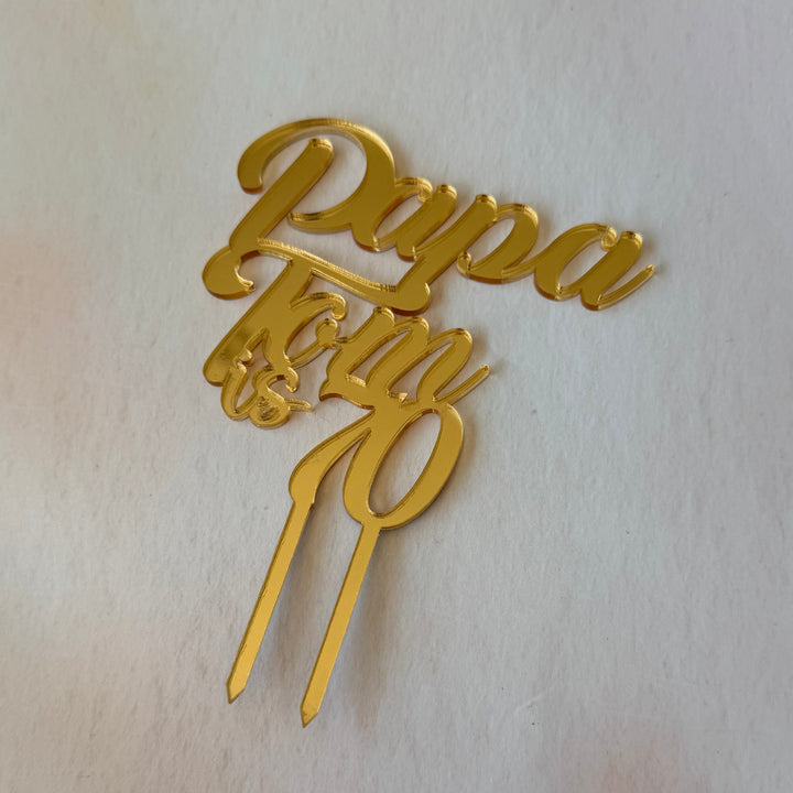 Gold metallic cake topper with the words 'Papa' and '10' in a script font, placed on a light beige textured surface.