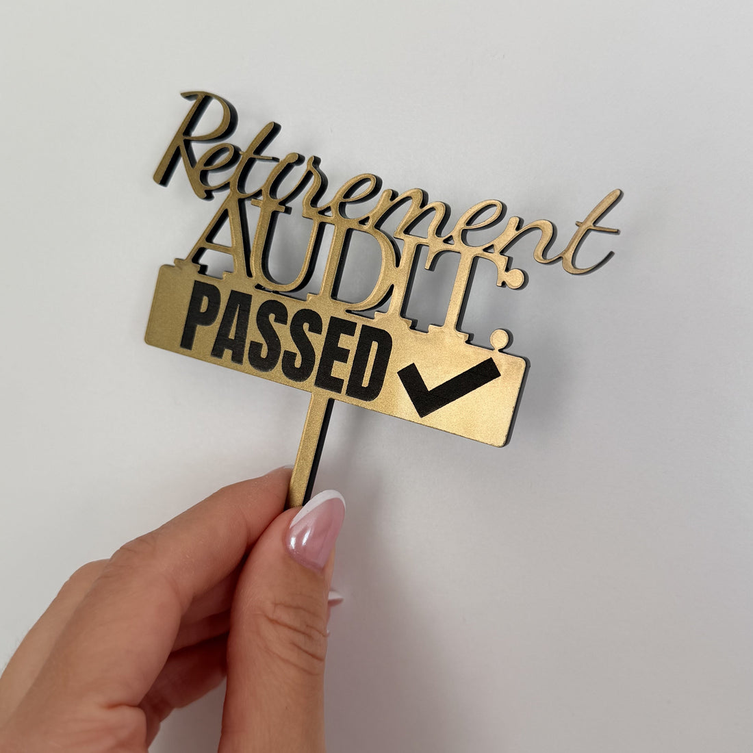 A hand with pink manicured nails holds a gold-colored wooden sign that reads 'Retirement AUDIT PASSED' with a checkmark, against a plain light gray background.