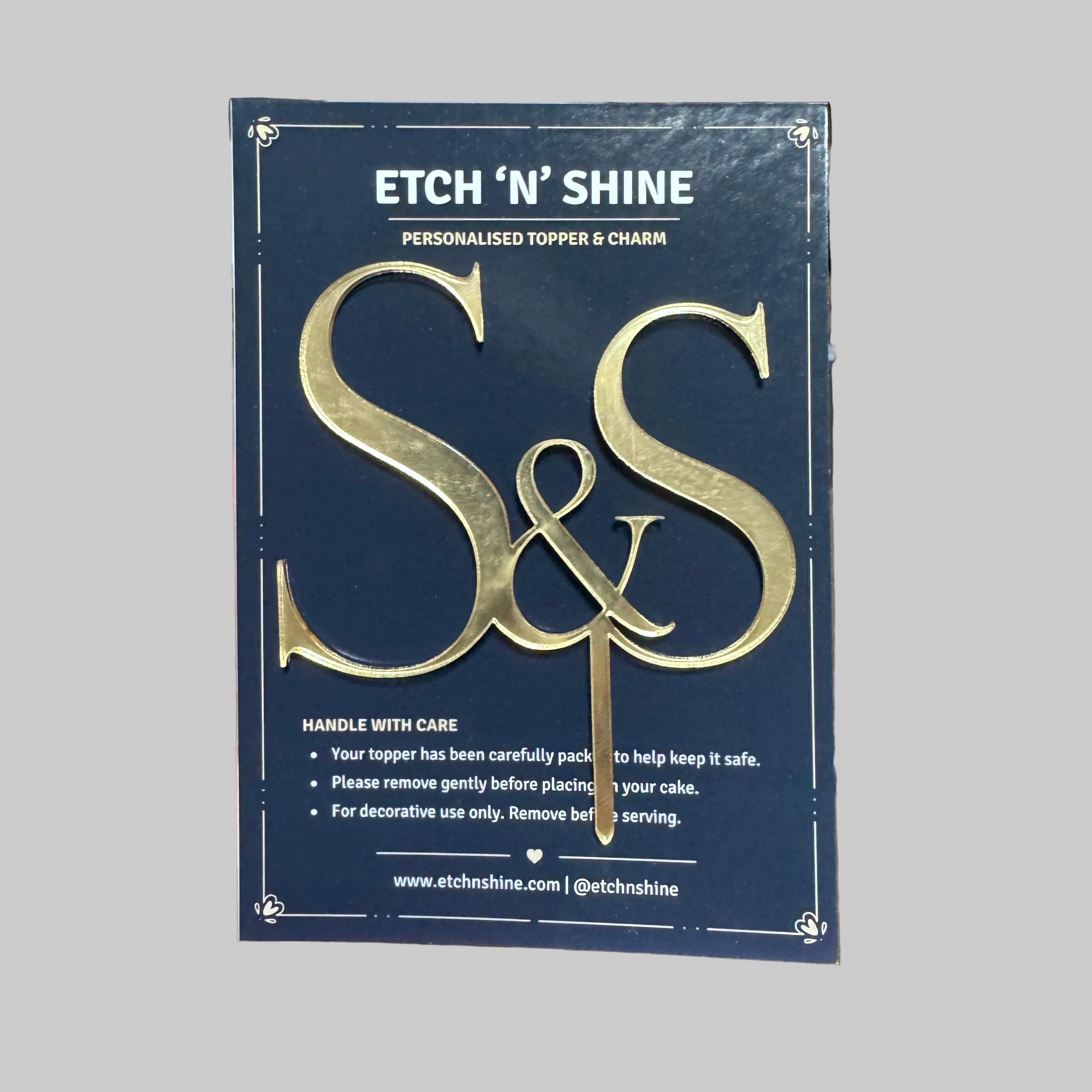 A hand holding a dark blue card for an 'Etch 'N' Shine' personalized topper and charm. The card features a large gold foil 'S&S' monogram and product details in white text.