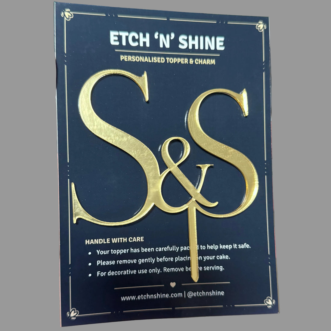 A hand holds a dark blue card backing for a custom gold 'S&S' topper and charm set by ETCH 'N SHINE. The backing card features elegant gold foil lettering and product details.