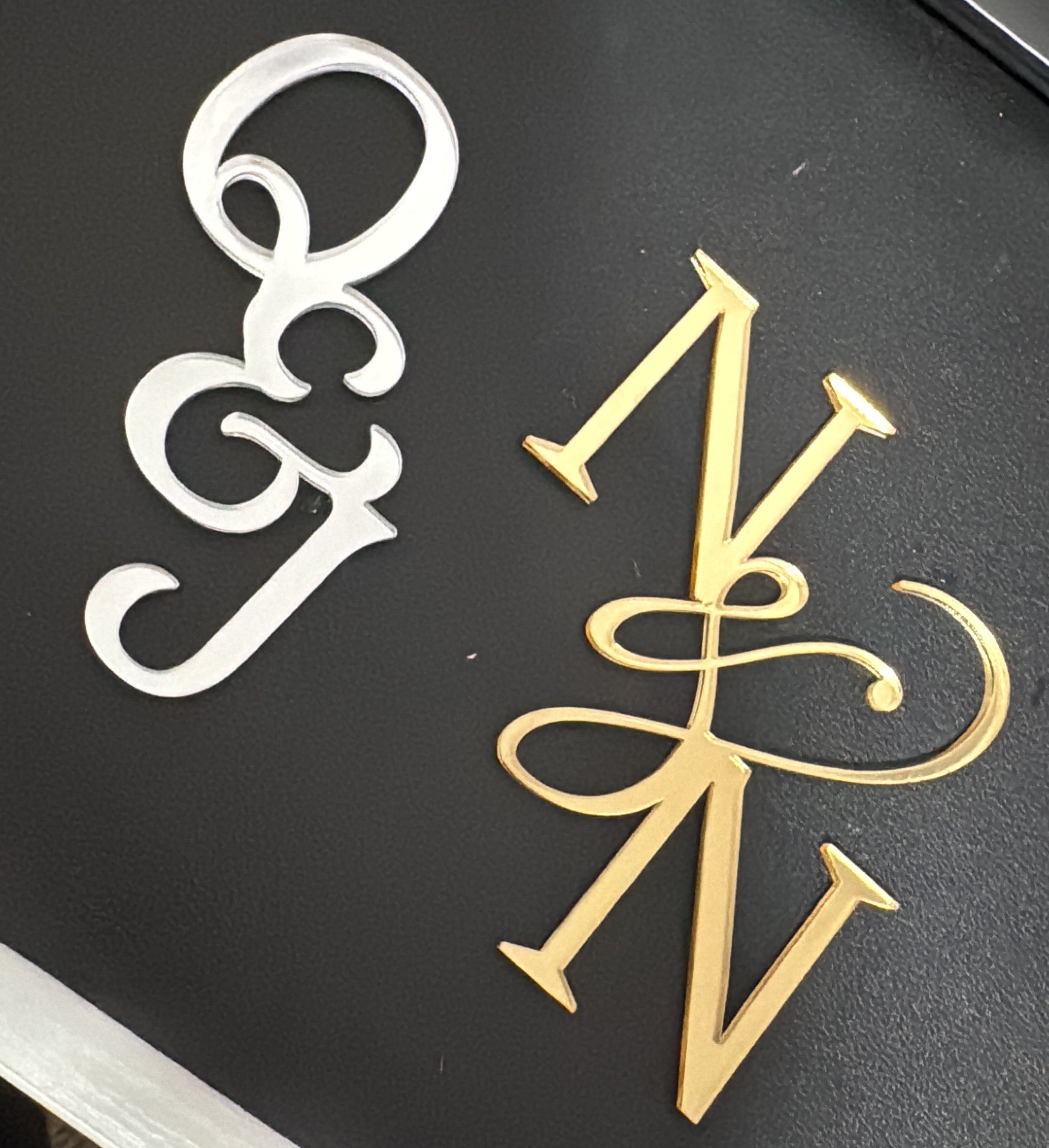 Two decorative monogram letter pieces, one white and one gold, displayed on a black textured surface.