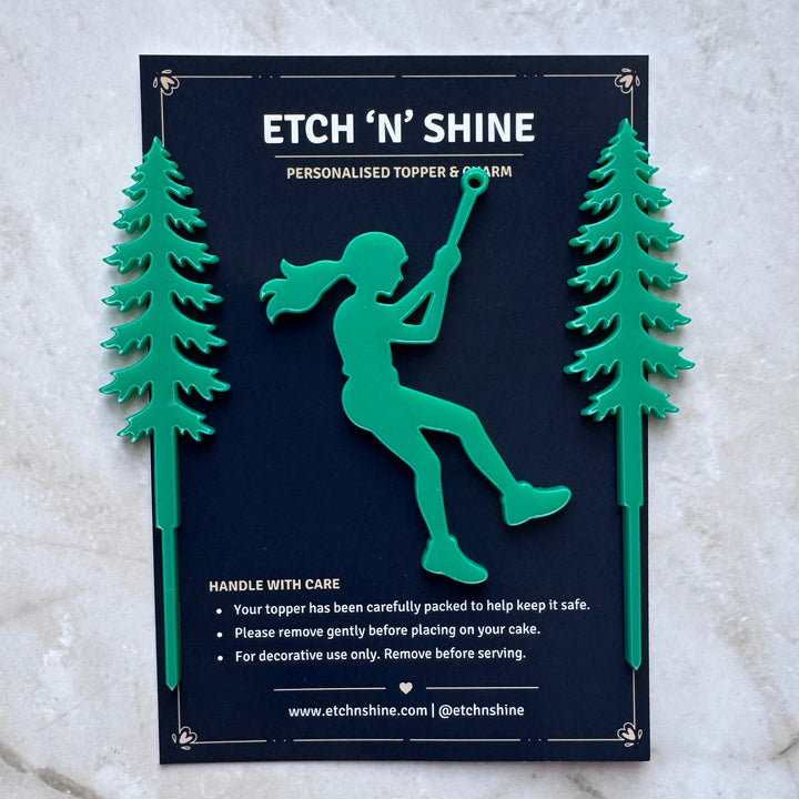 A personalized wooden topper and bark mount plaque reading 'ETCH 'N SHINE' with a dark navy center, green silhouette of a person climbing a tree, and two green pine trees on either side, displayed on a light marble surface.
