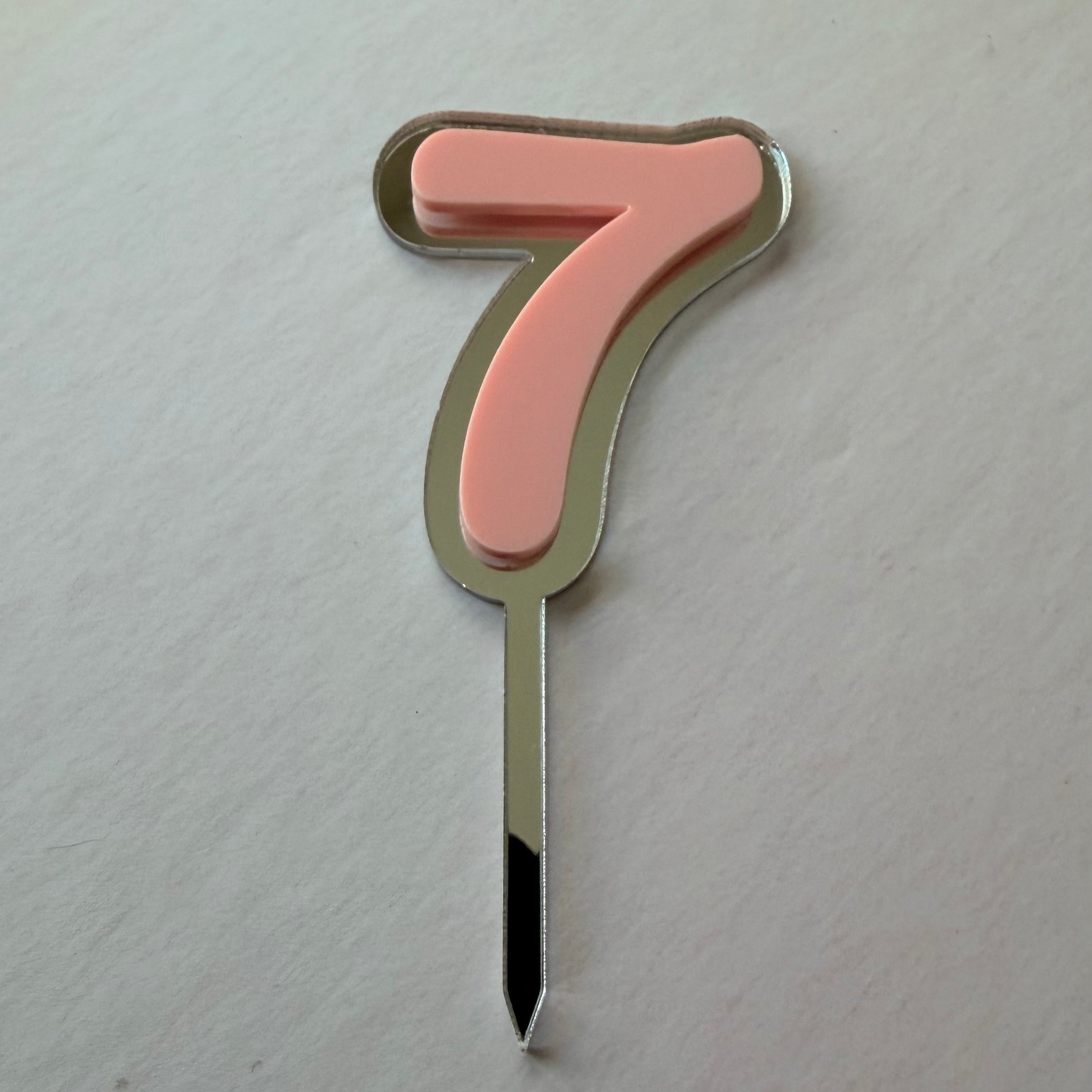 A single number 7 birthday candle with a pastel pink enamel face, a gold-toned metal border, and a silver-toned metal stem, photographed on a light off-white textured background.