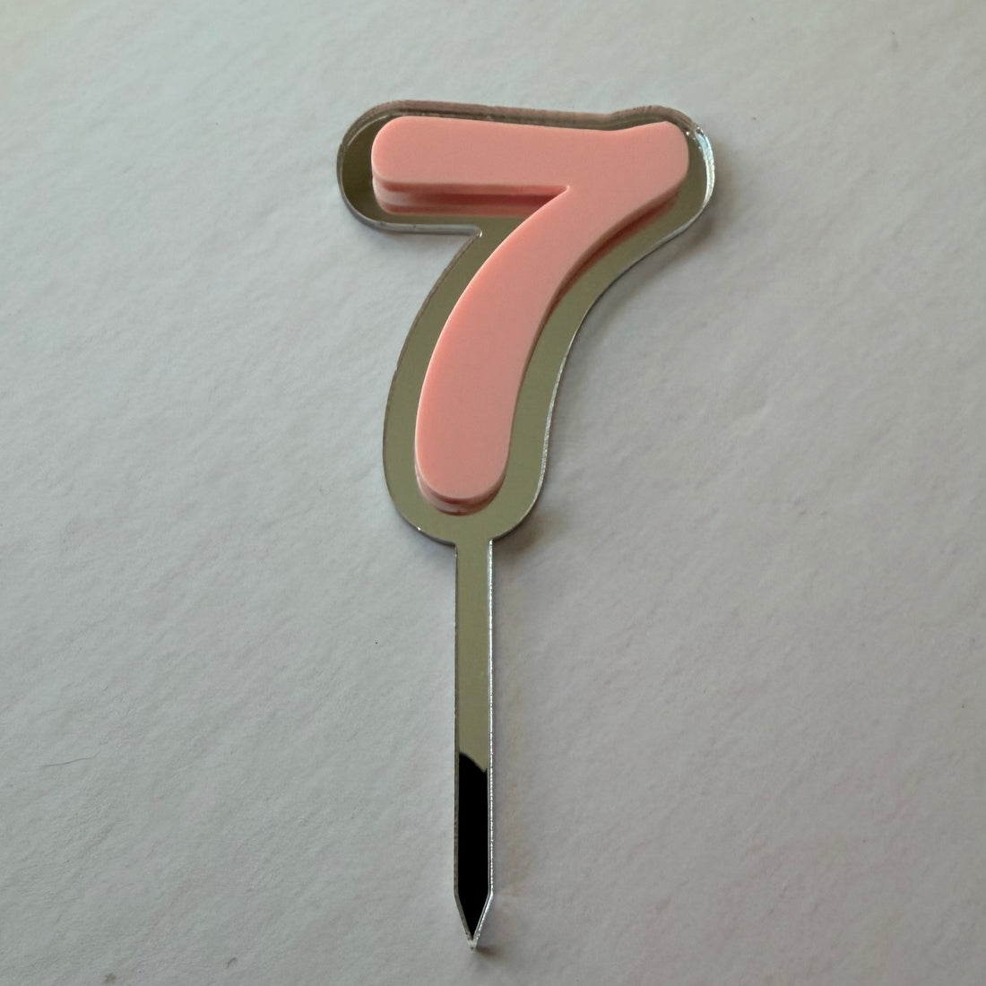 A single number 7 birthday candle with a pastel pink enamel face, a gold-toned metal border, and a silver-toned metal stem, photographed on a light off-white textured background.