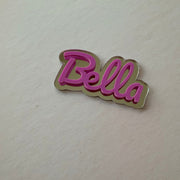 A small metallic pin or charm with the word 'Bella' in pink script on a gold-toned outline, placed on a plain off-white surface.