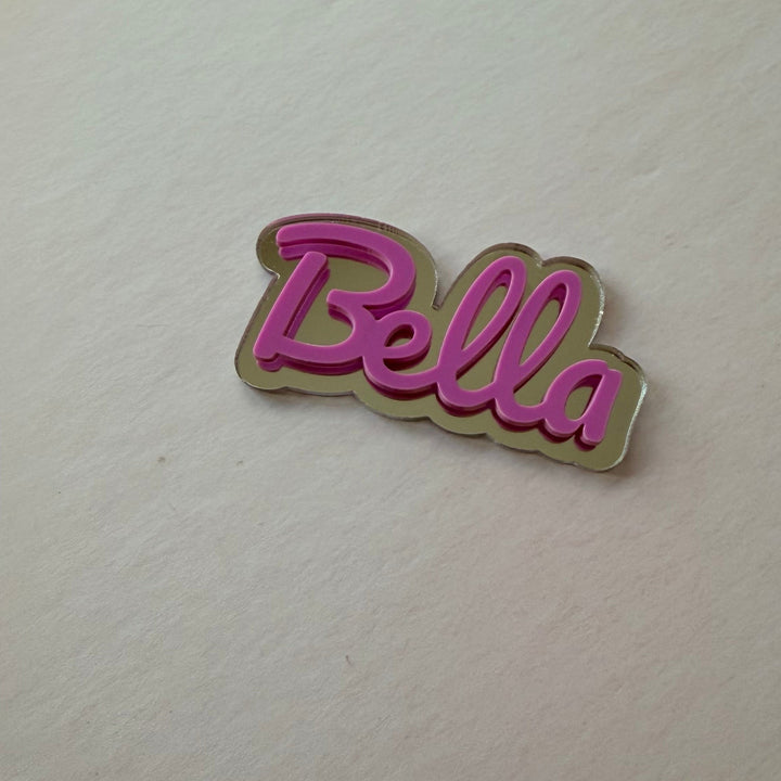 A small metallic pin or charm with the word 'Bella' in pink script on a gold-toned outline, placed on a plain off-white surface.