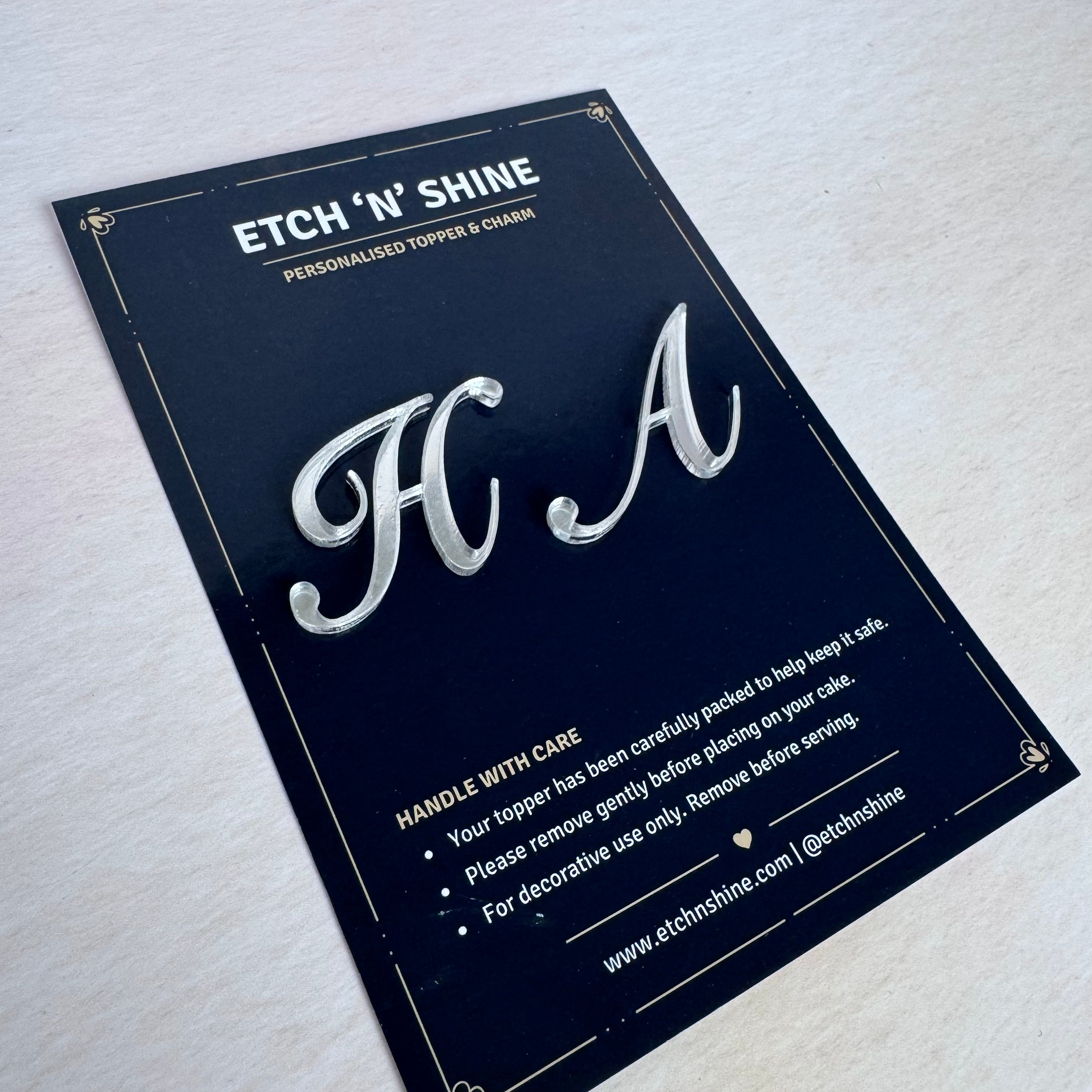 A close-up of a silver metallic monogram charm shaped like the letters 'HA', mounted on a dark navy blue display card. The card features gold borders, white text, and care instructions, and is photographed on a light-colored wooden surface.