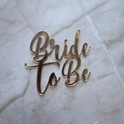 Gold metallic script lettering reading 'Bride to Be' placed on a light marble surface.