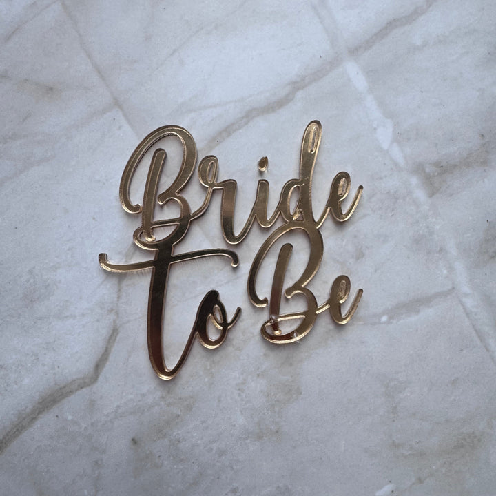 Gold metallic script lettering reading 'Bride to Be' placed on a light marble surface.
