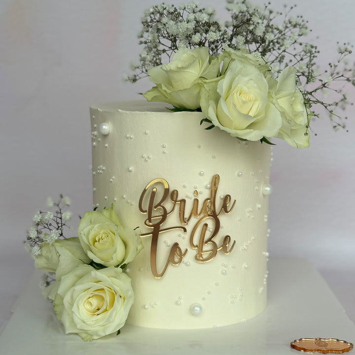 A frosted wedding cake with white roses and baby's breath, featuring a gold 'Bride To Be' script topper on a white board.