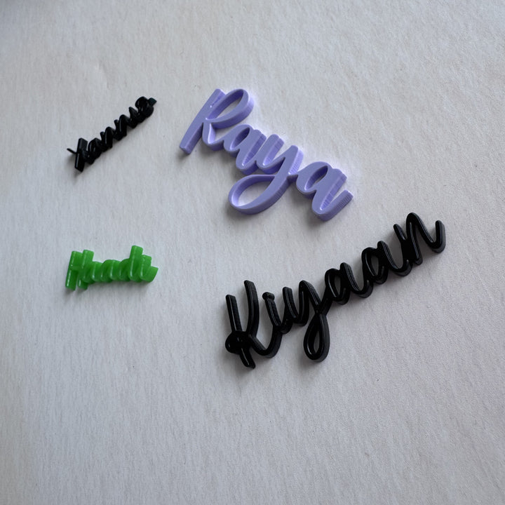 Four cursive nameplates or decals in black and lavender, with one small green one, arranged on a textured off-white background.