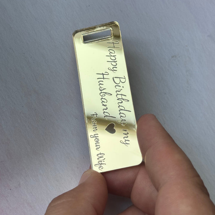 A hand holding a small rectangular gold-toned metal card or tag with engraved text reading 'Happy Birthday my Husband' and 'you are my life' on a light surface.