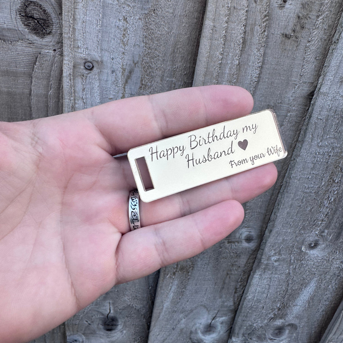 A hand holding a personalized engraved metal keychain that reads 'Happy Birthday my Husband From your Wife' against a gray wooden fence background.