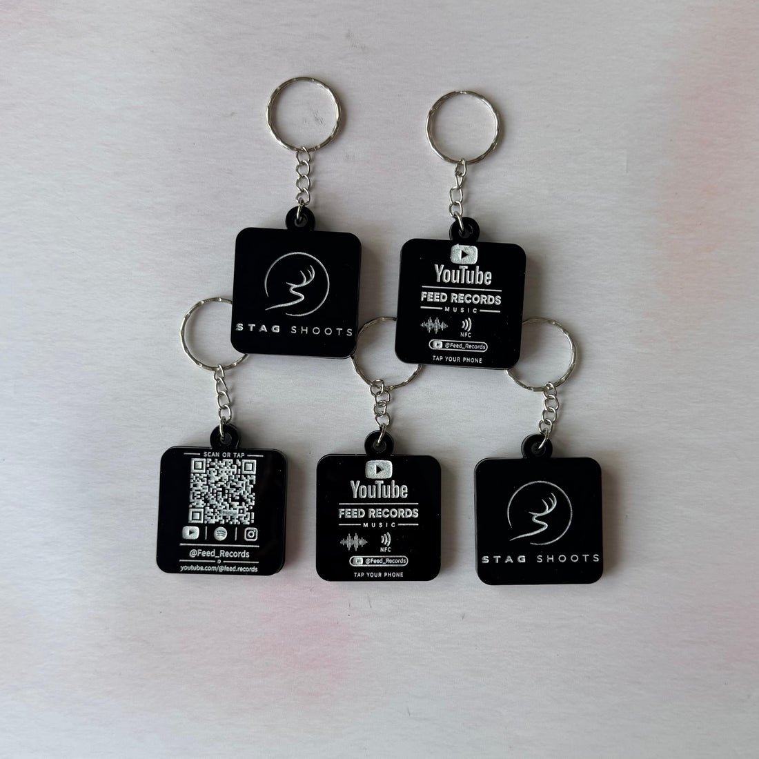 Five black square keychains with silver metal rings, arranged on a light off-white surface. Each keychain has a small rectangular black tag printed with white text and logos, including 'YouTube', 'FRED RECOORDS', and 'STAR SHOOTS'.