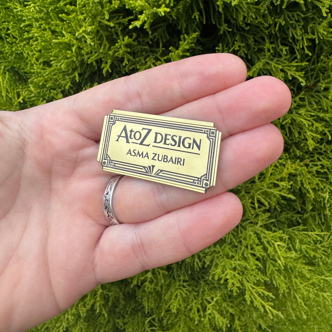 Hand holding a gold and black business card with 'AtoZ Design' branding against a green leafy background.