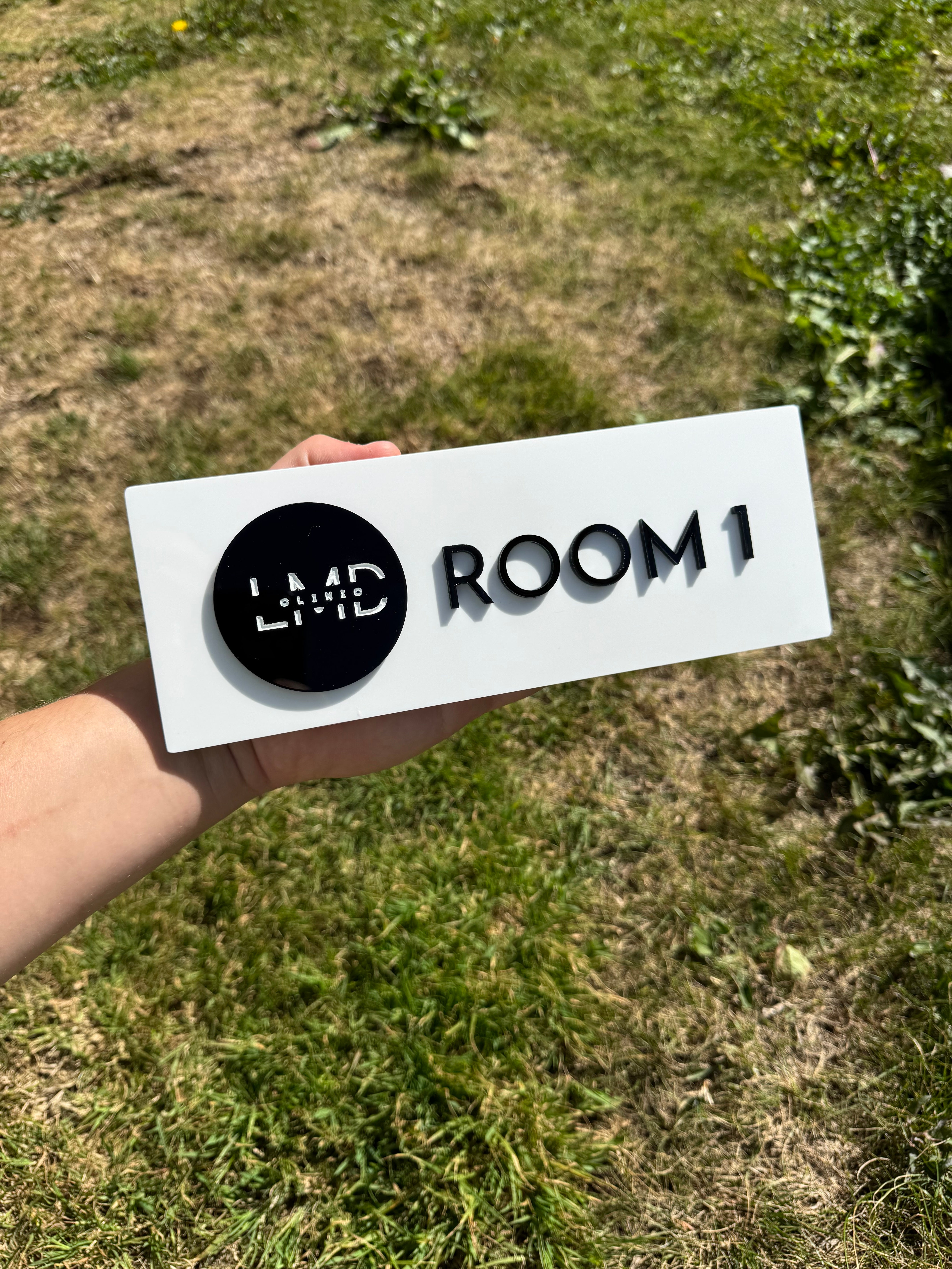 Hand holding a white sign with 'ROOM 1' and 'LMD' printed on it against a grassy background.