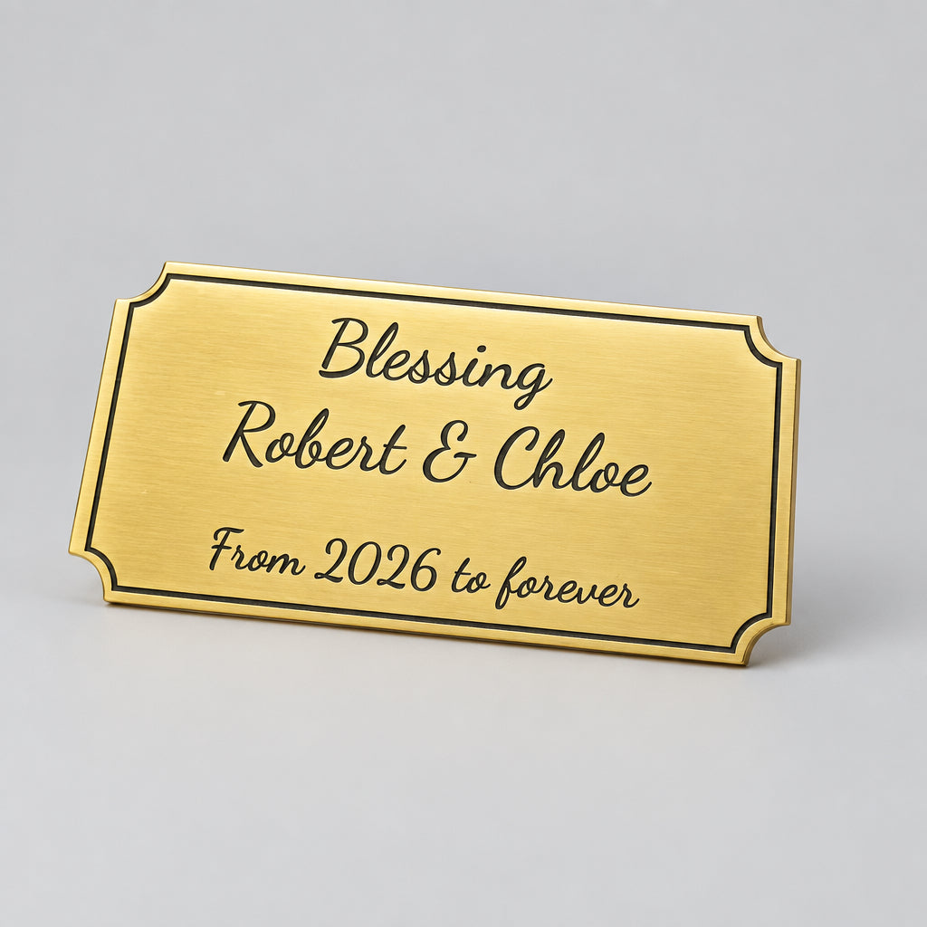 A rectangular gold keepsake plaque engraved with 'Blessing Robert & Chloe From 2026 to forever' resting on a dark textured surface near a keyboard.