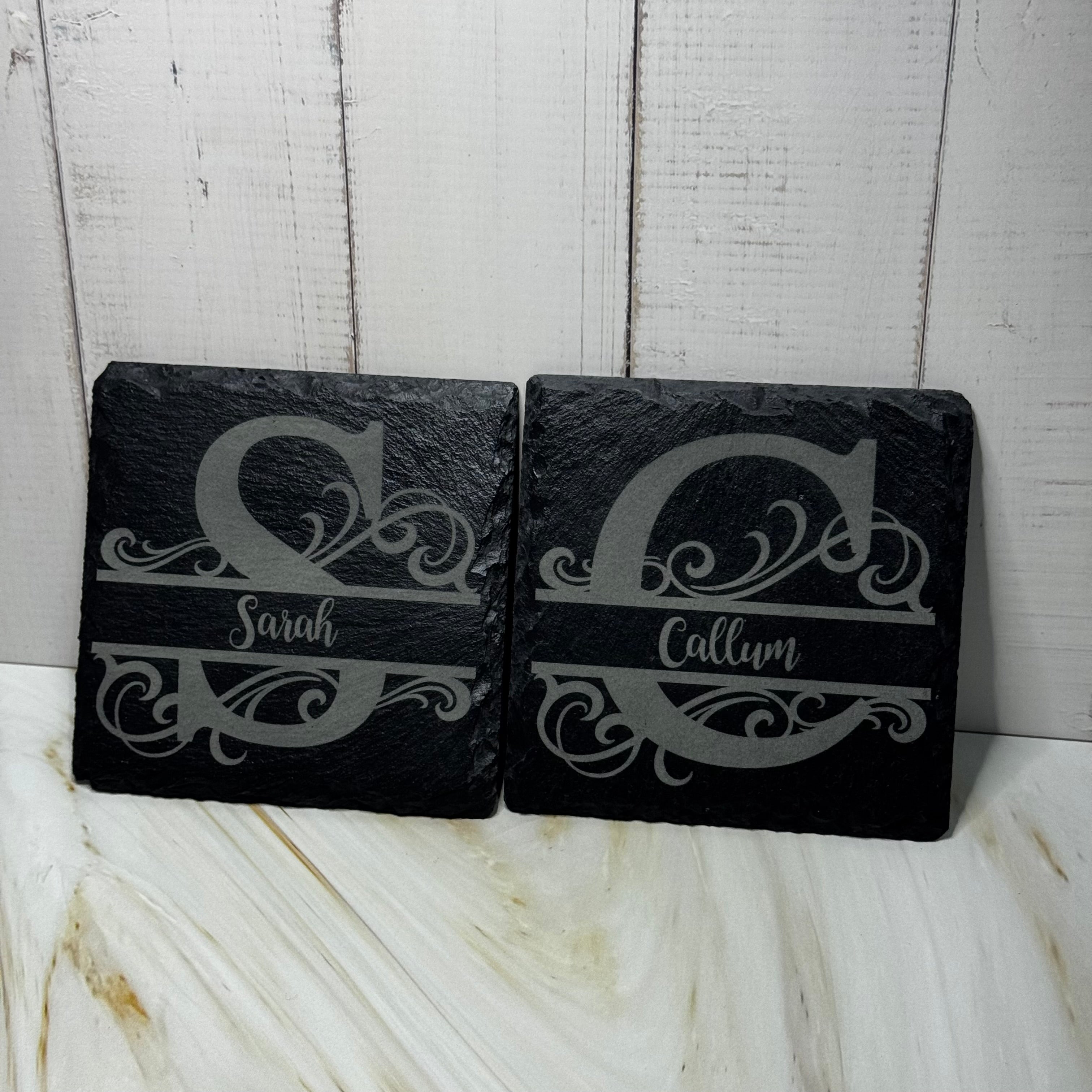 Two black slate coasters with engraved monograms on a light wooden surface.