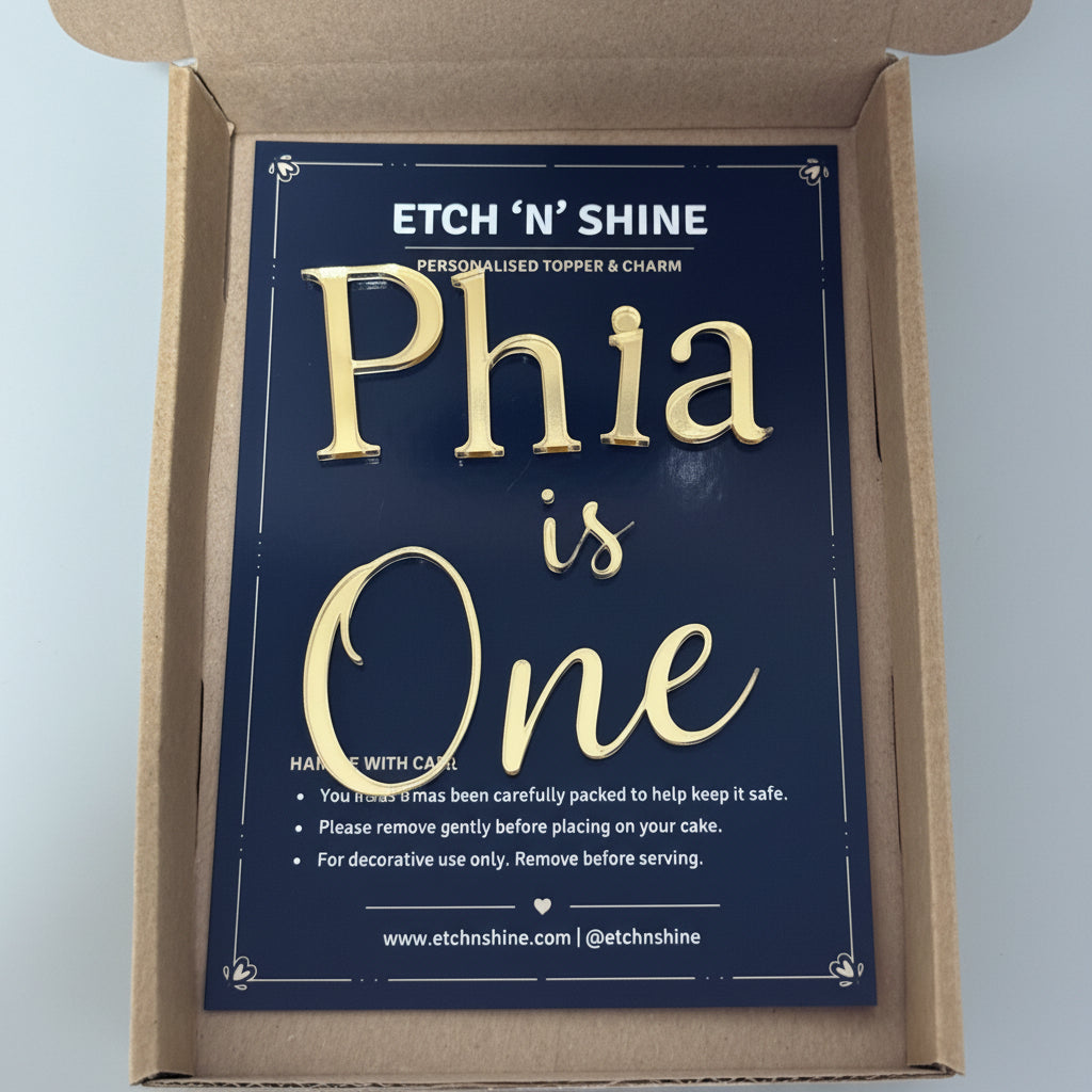 A navy blue personalized topper and charm card with gold lettering reading 'Phia is One', displayed inside an open brown cardboard box. The brand name 'ETCH 'N' SHINE' is visible at the top of the card.