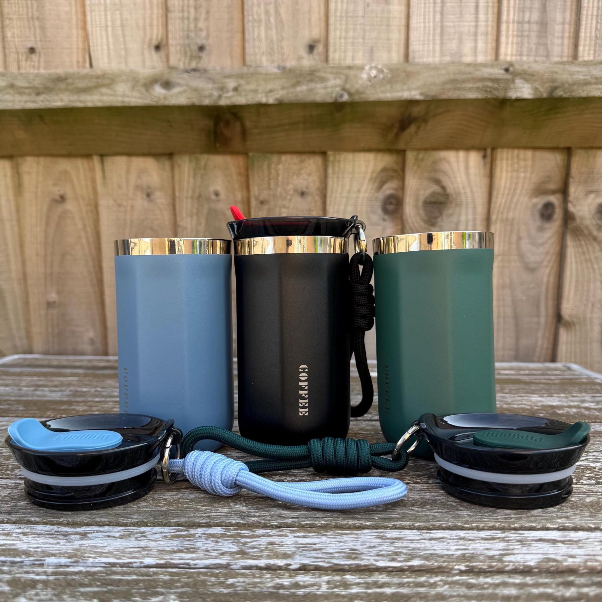 Set of three insulated stainless steel coffee mugs in black, blue, and green with matching rope handles and lids, custom name engraving available, perfect for gifting.