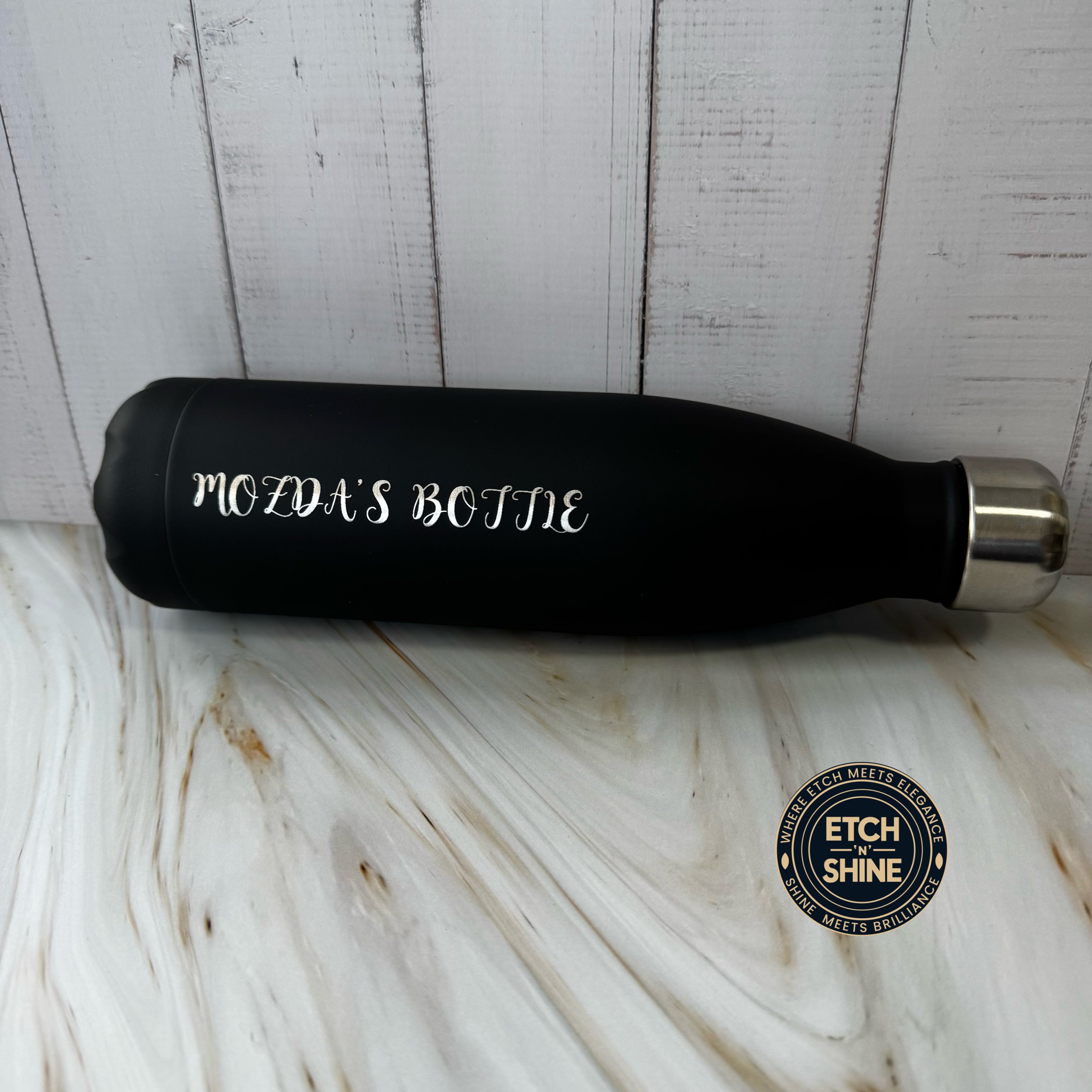 Black water bottle with 'MOZDA'S BOTTLE' text on a white marble surface, branded 'ETCH SHINE'.