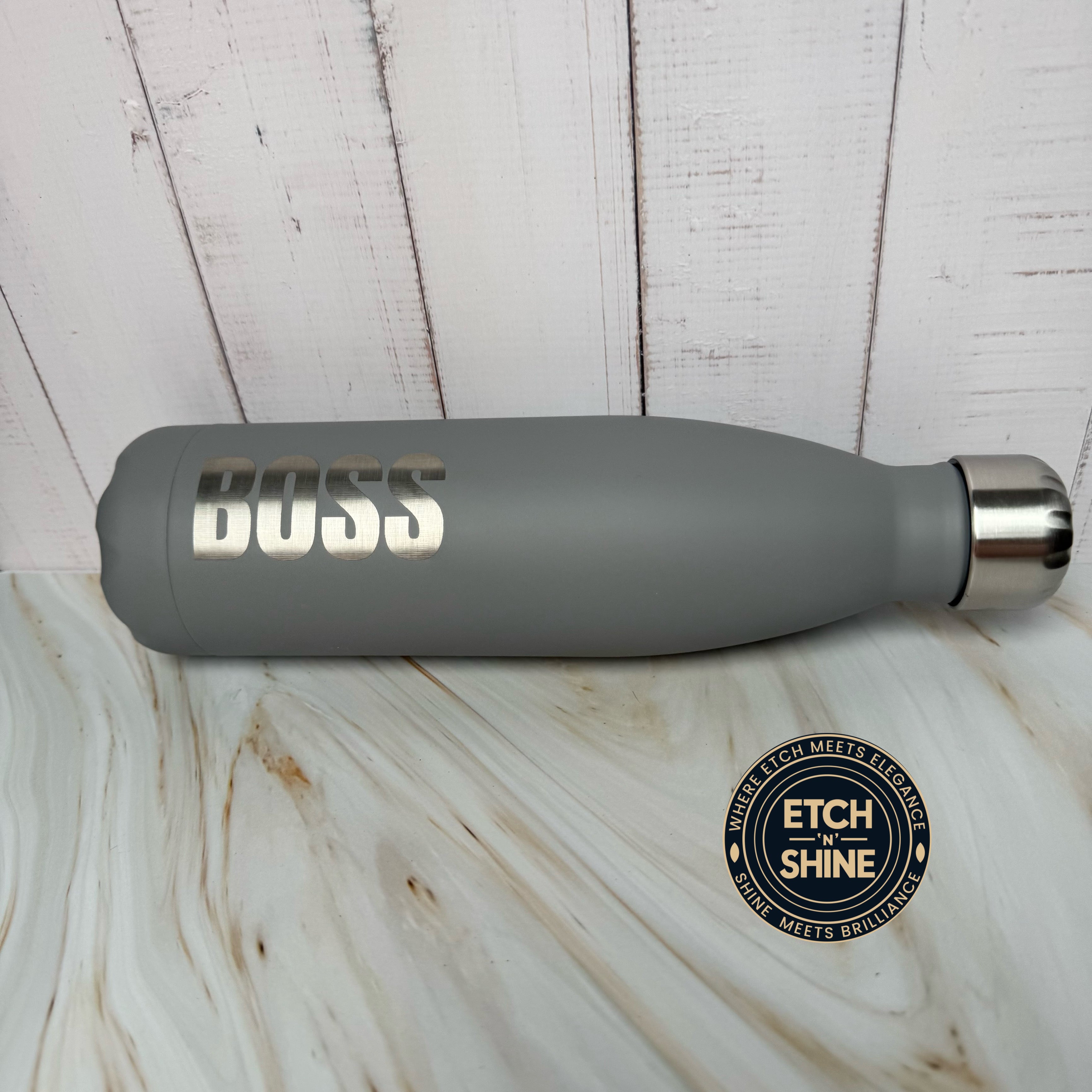 Gray water bottle with 'DUSS' branding on a wooden surface, featuring Etch 'N Shine logo.