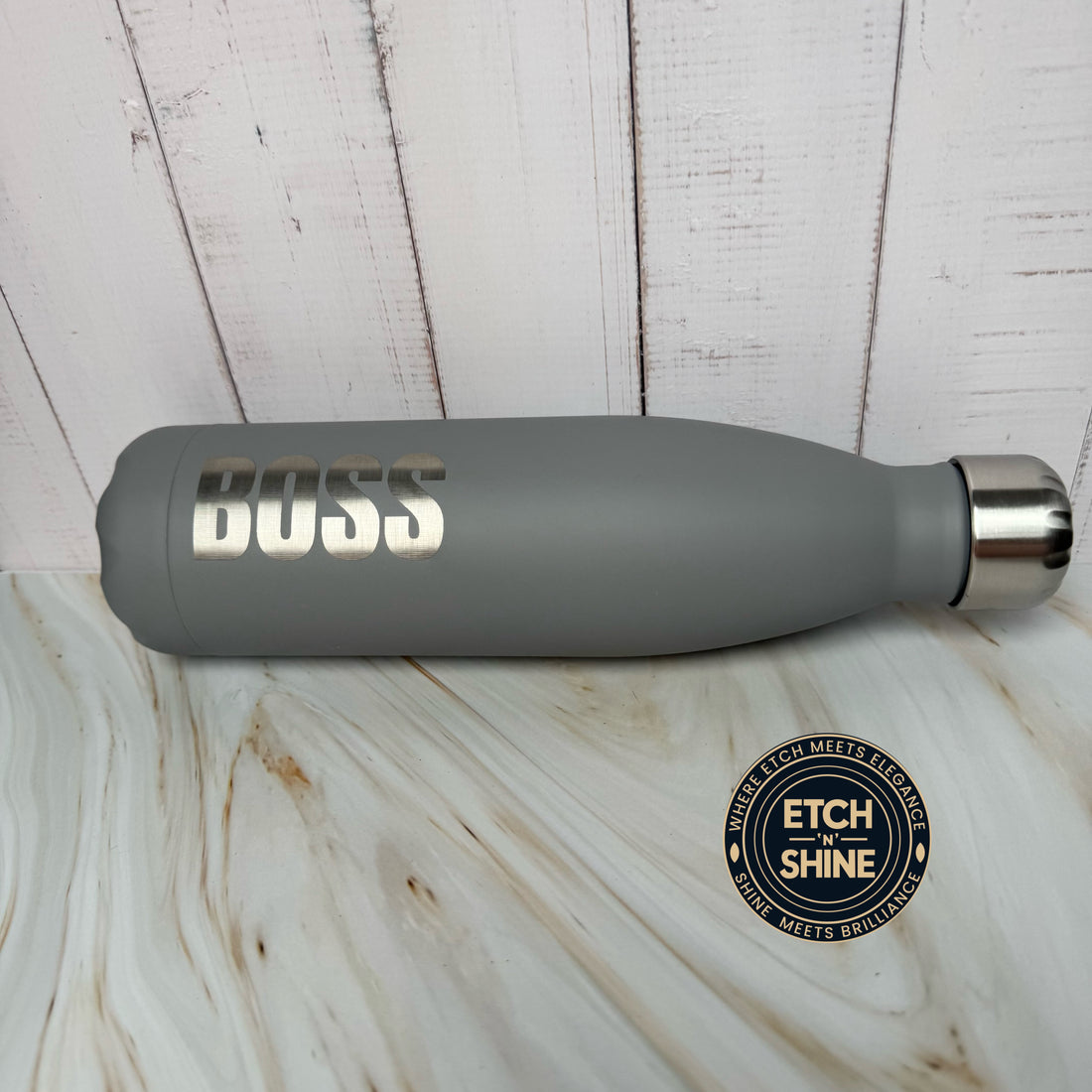 Gray water bottle with 'DUSS' branding on a wooden surface, featuring Etch 'N Shine logo.