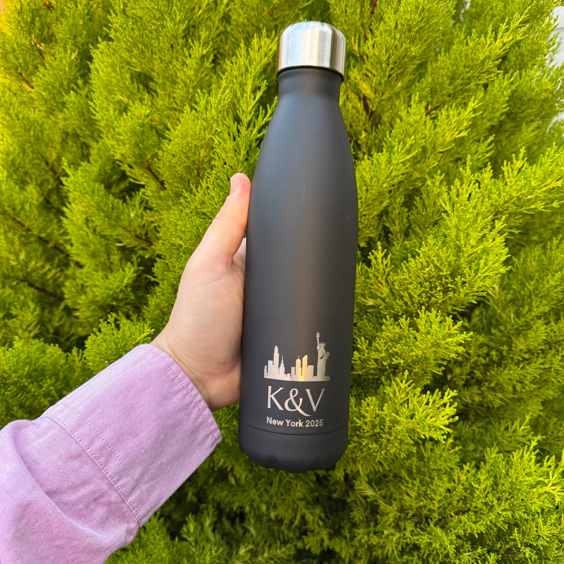 Person holding a black water bottle with 'K&V New York 2023' design against a green bush background
