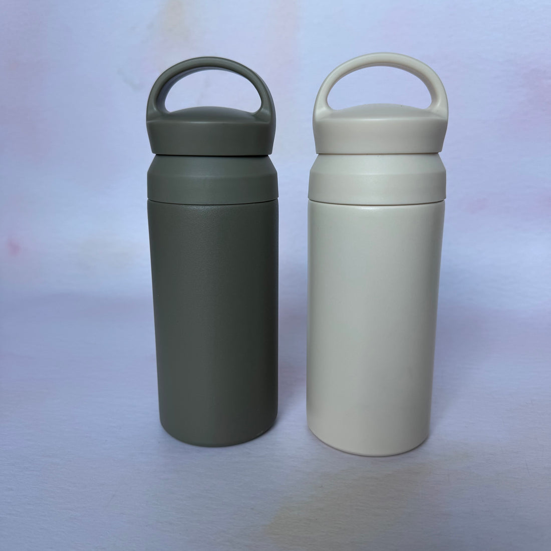 Two insulated travel mugs, one green and one beige, on a light purple background.