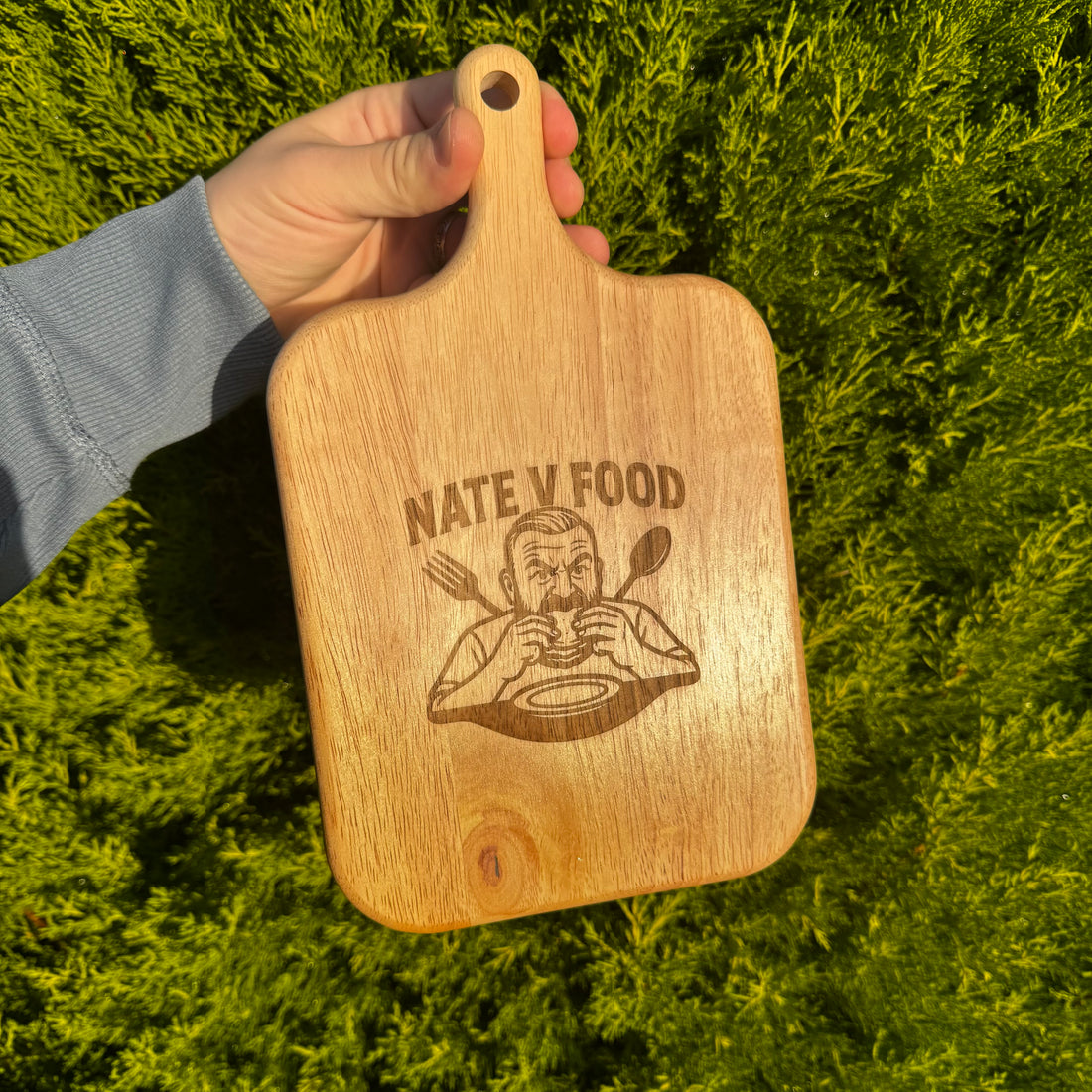 Wooden cutting board with 'Nate Y Food' engraving held in front of green foliage