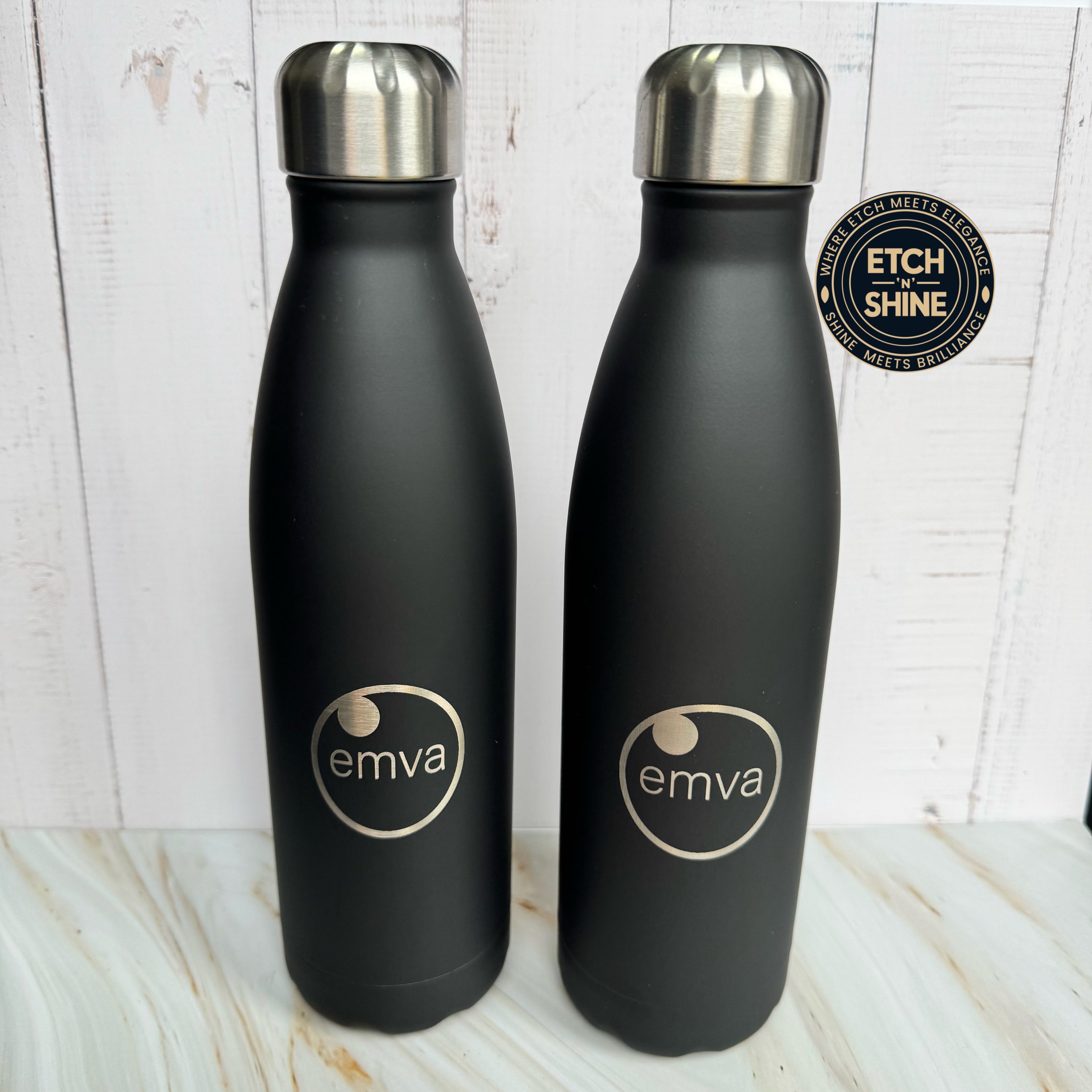 Two black water bottles with silver caps on a light wooden surface, featuring the 'emva' logo.