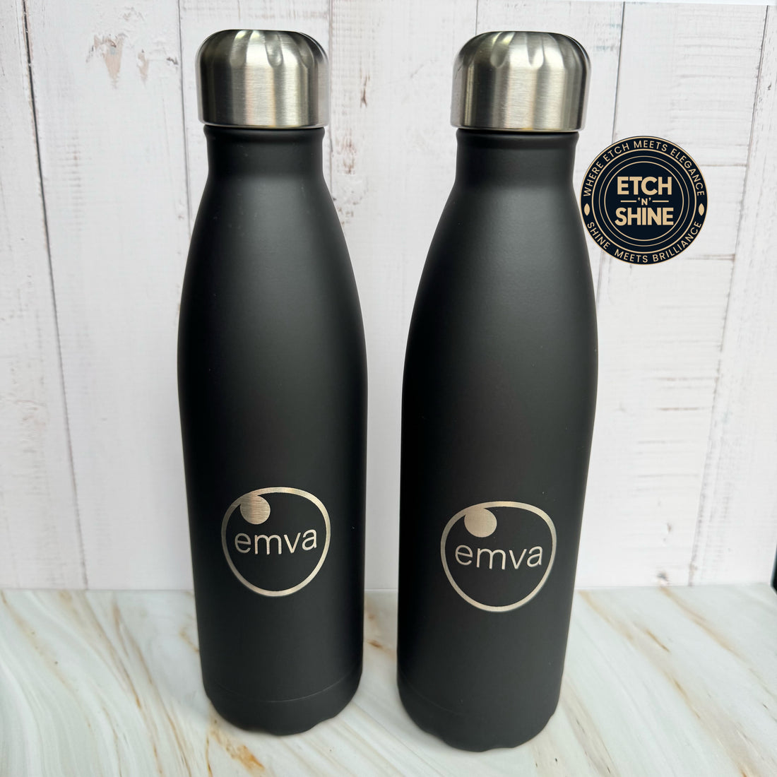 Two black water bottles with silver caps on a light wooden surface, featuring the 'emva' logo.