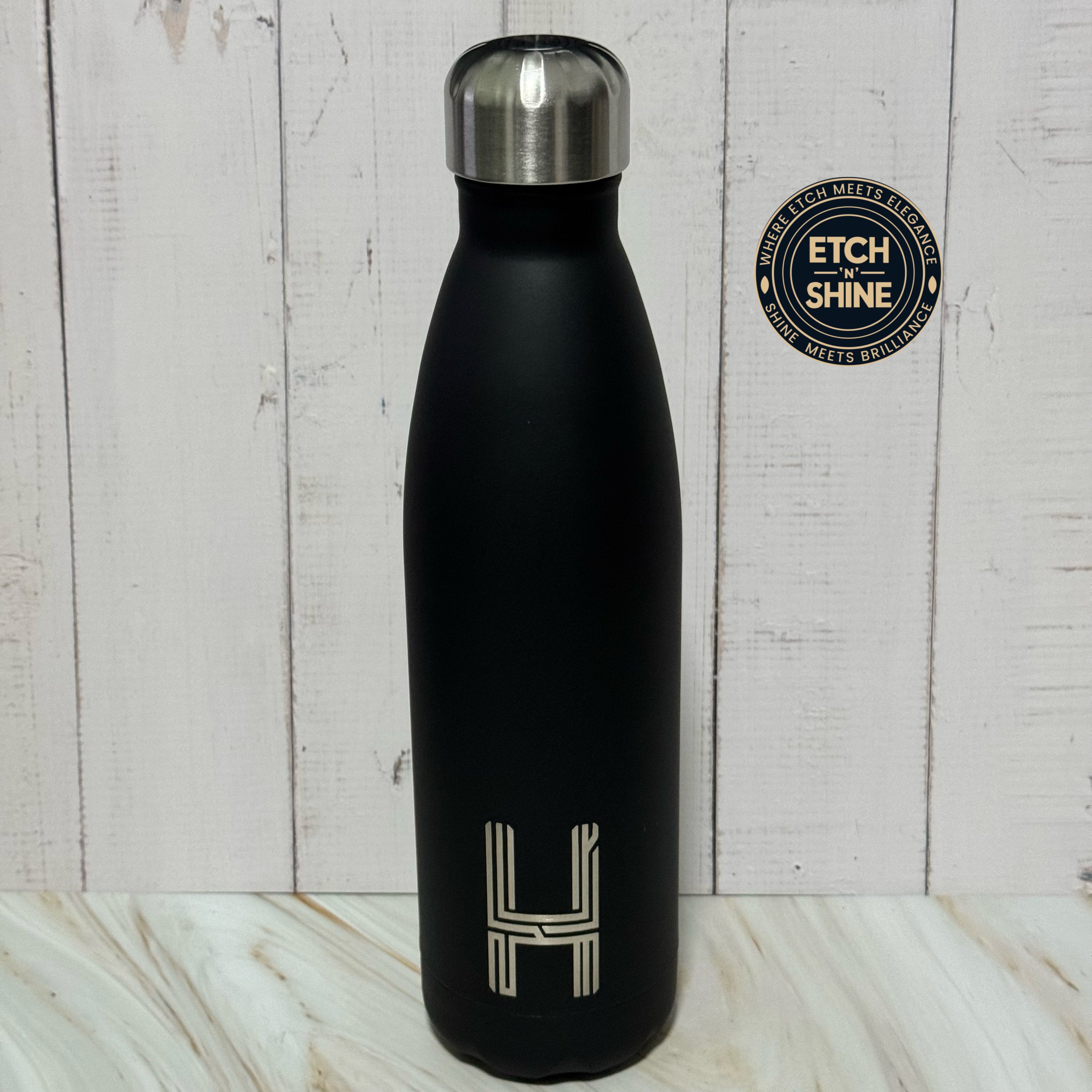 Black water bottle with silver cap on a wooden surface, featuring 'ETCH SHINE' branding.