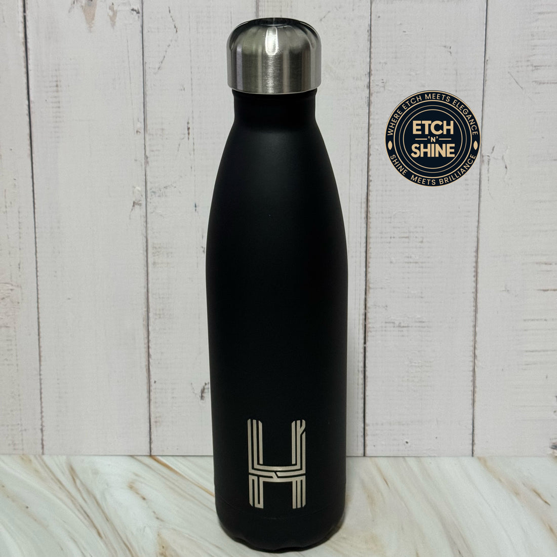 Black water bottle with silver cap on a wooden surface, featuring 'ETCH SHINE' branding.