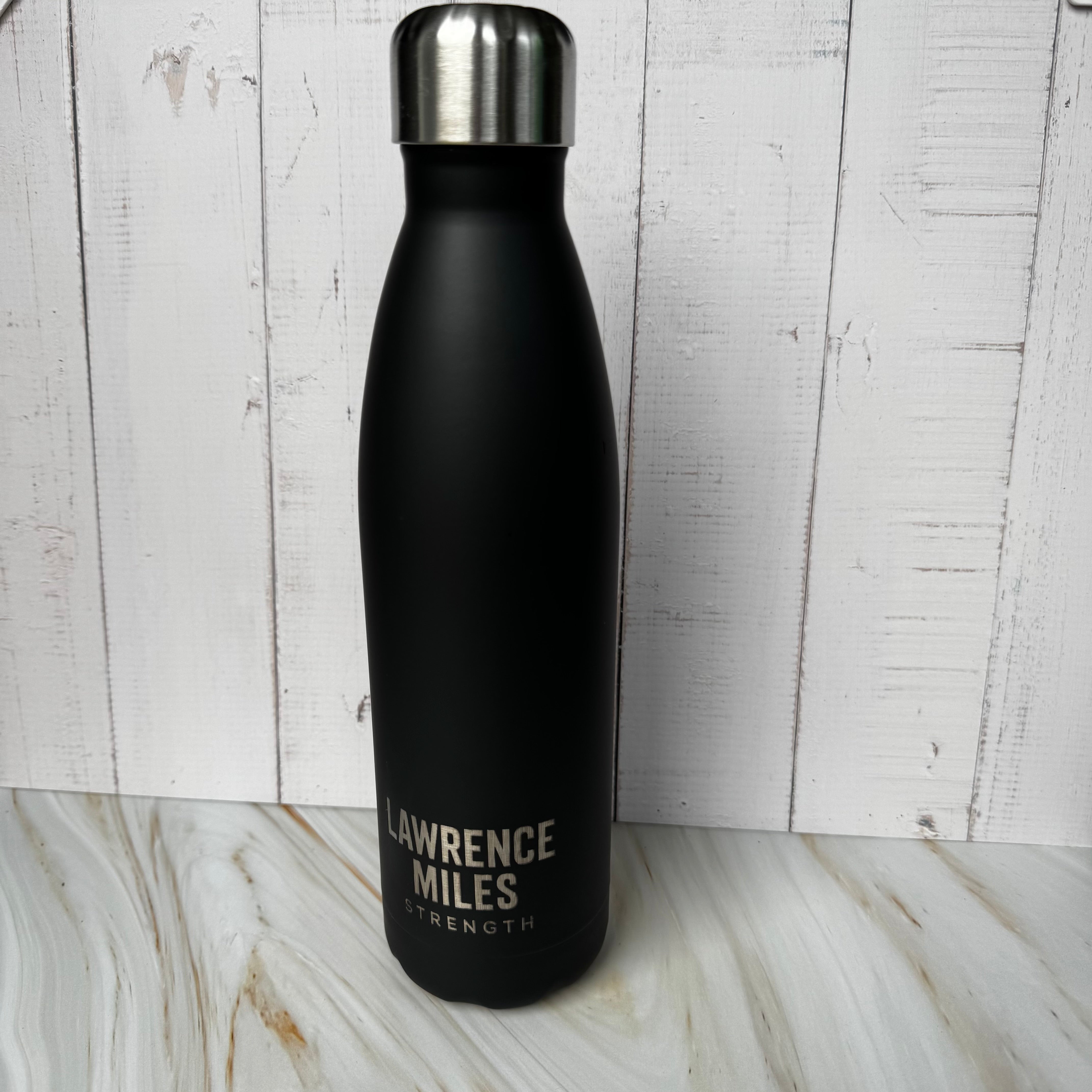Black water bottle with 'Lawrence Miles Strength' text on a wooden surface