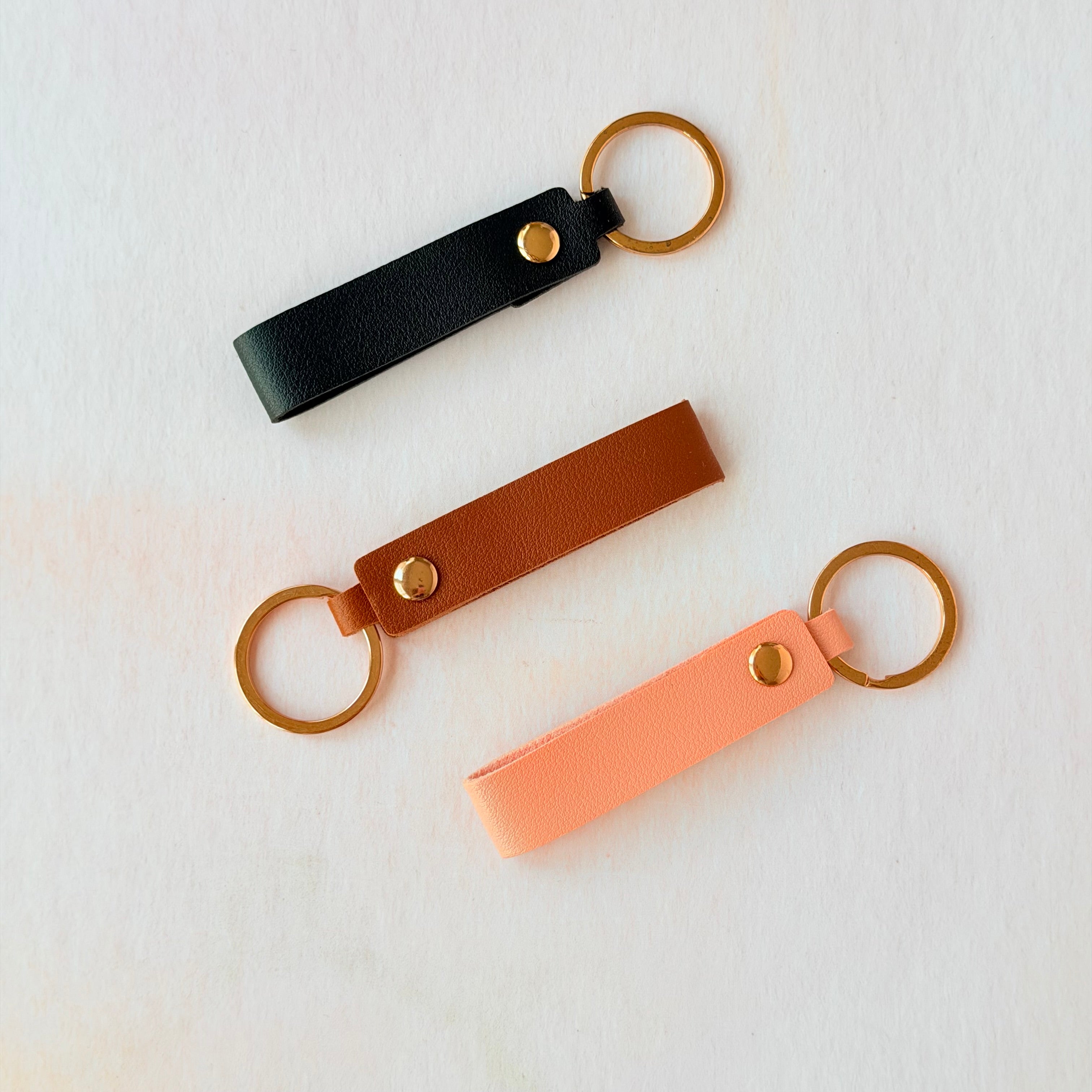 Three leather keychains in black, brown, and pink on a light gray background