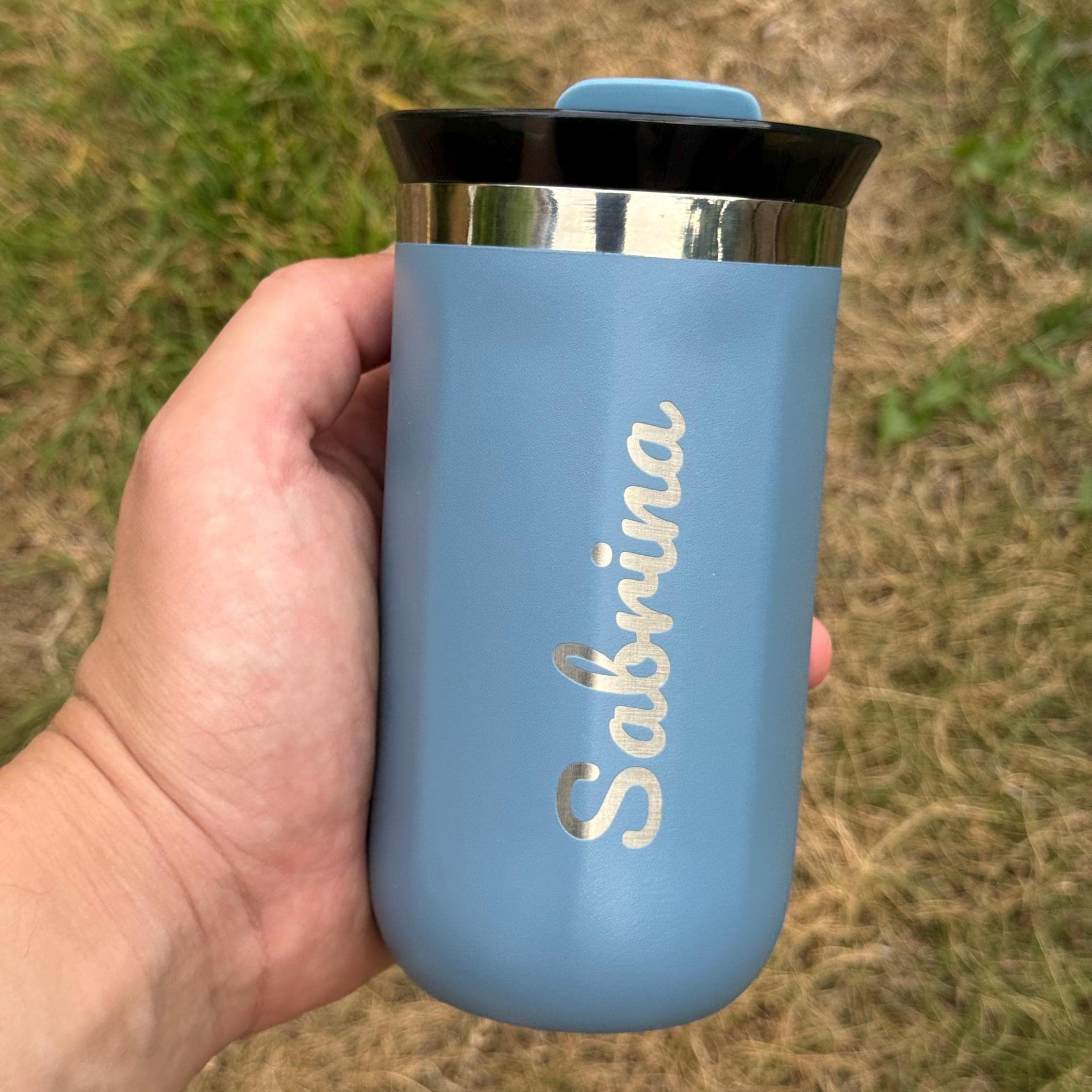 Blue stainless steel travel cup with silver engraved name “Sabrina” in script font. Features black sipping lid and metallic rim. Compact and stylish, perfect for personalized gifting or daily coffee use.