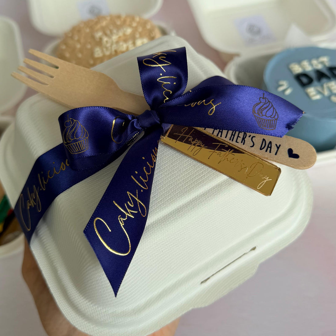 Hand holding a cake with a blue ribbon and 'Happy Father's Day' mirror acrylic tag, with a blurred background.