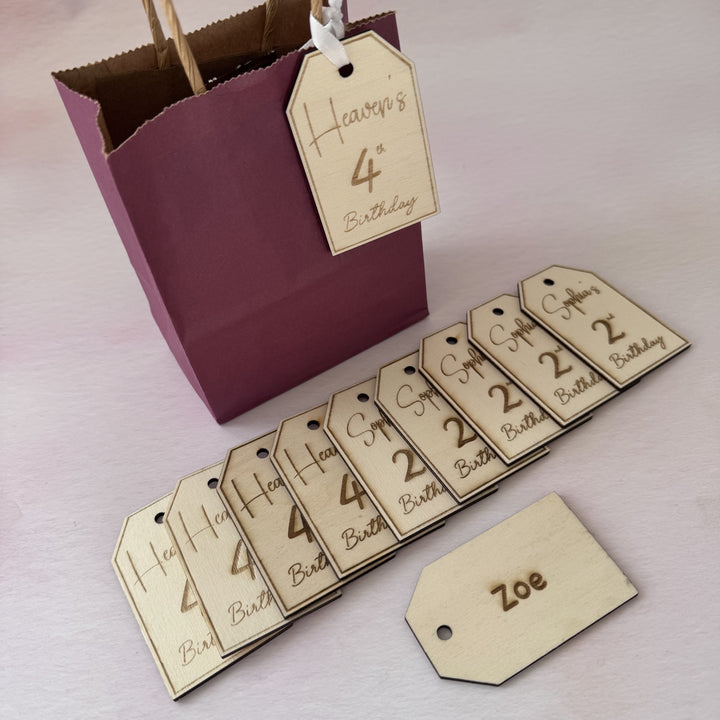 Set of wooden tags with numbers and names, including a maroon gift bag with a tag, on a light pink background.